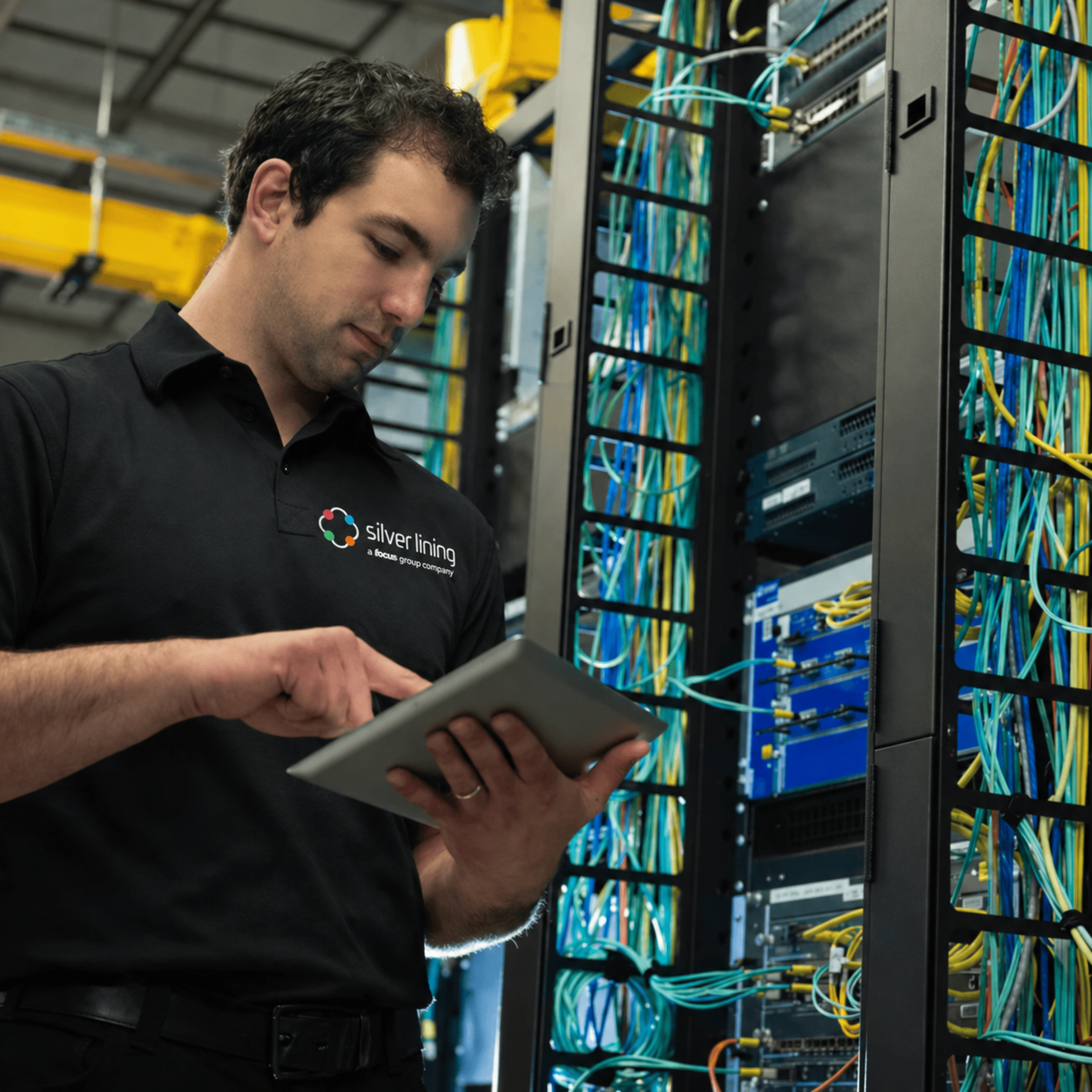 Network engineer in a “Silver Lining” polo using a tablet beside server racks and patch panels, configuring switching and routing infrastructure for secure, high-performance data networks.