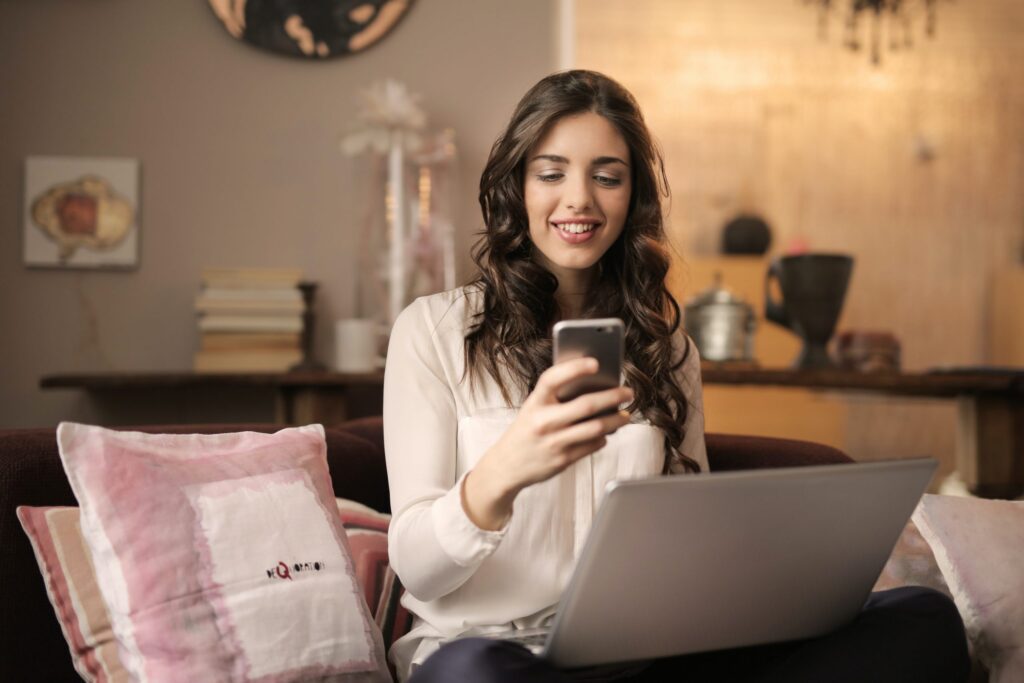 Woman sitting on a sofa using a smartphone and laptop.