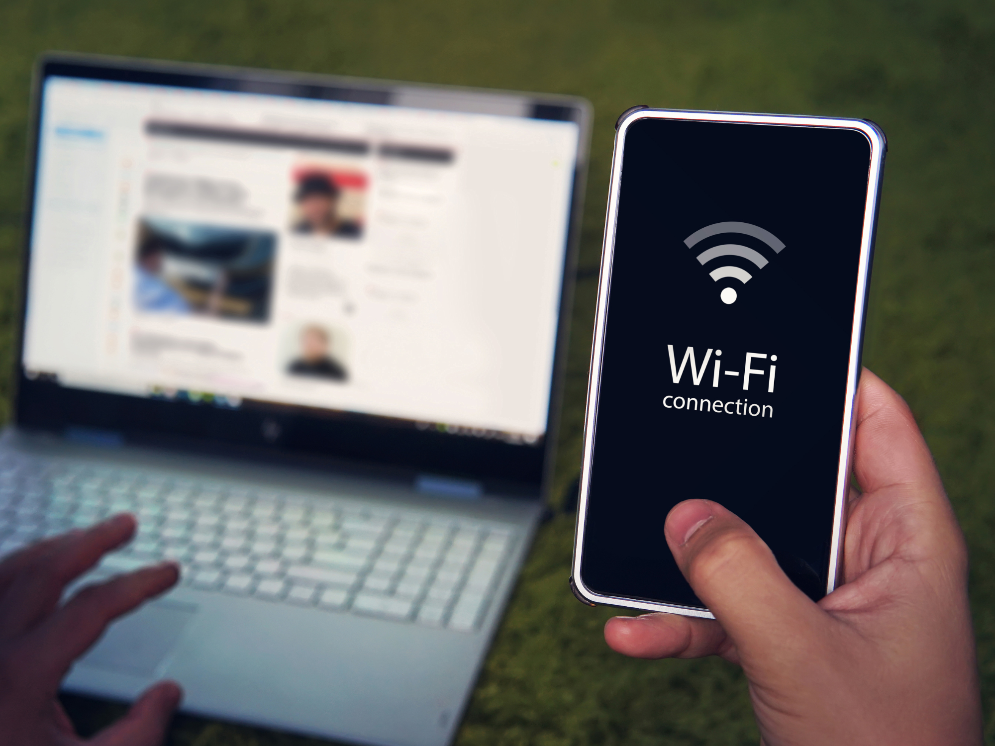 Person holding a smartphone displaying a Wi-Fi connection symbol beside an open laptop outdoors.