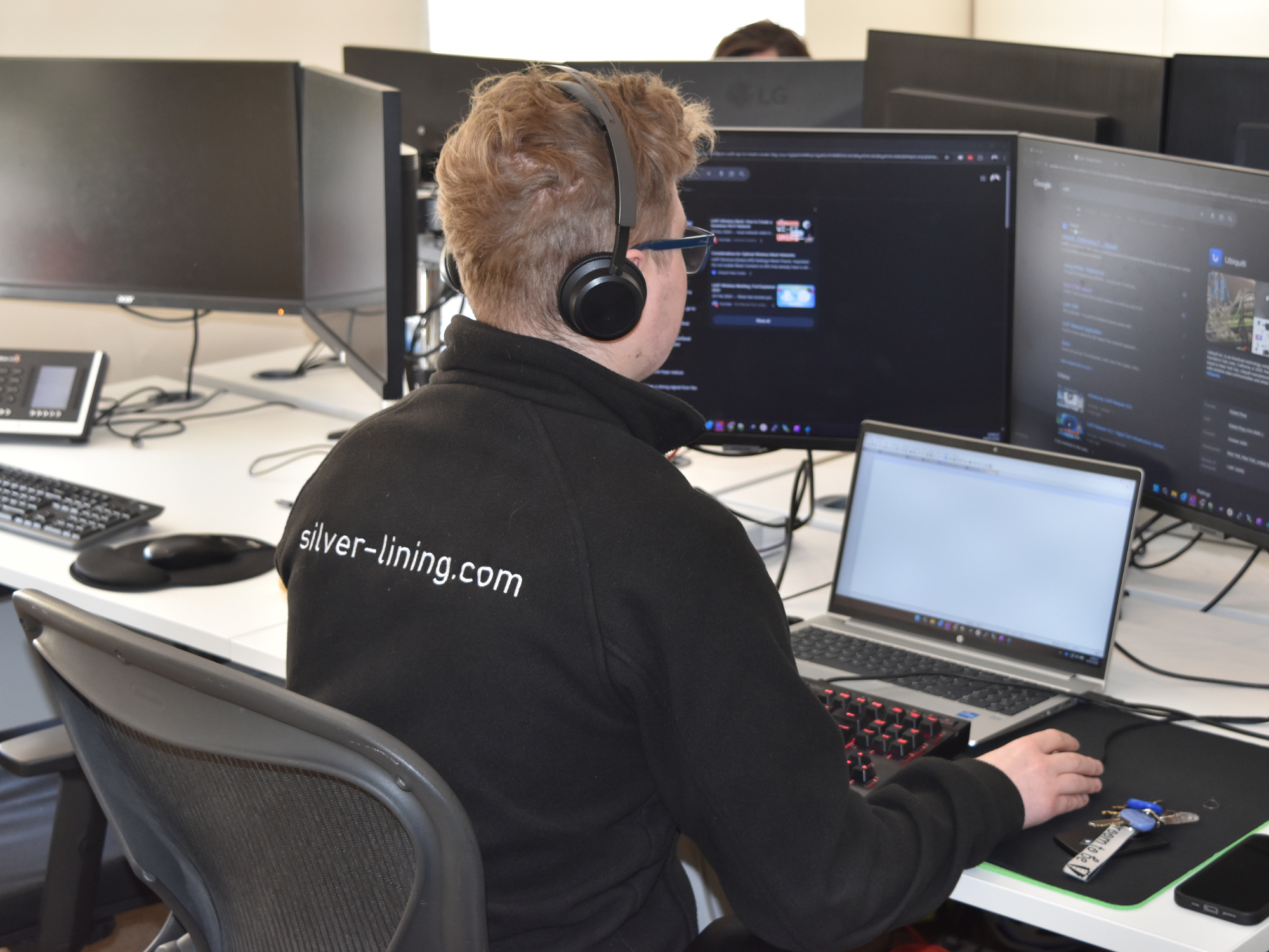 An IT support engineer wearing a headset sits at a desk, working on a laptop and multiple monitors to provide managed IT services.